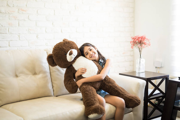 fröhliches kleines Mädchen umarmt Spielzeugbär und lächelt, während es zu Hause auf dem Sofa sitzt  - Foto, Bild