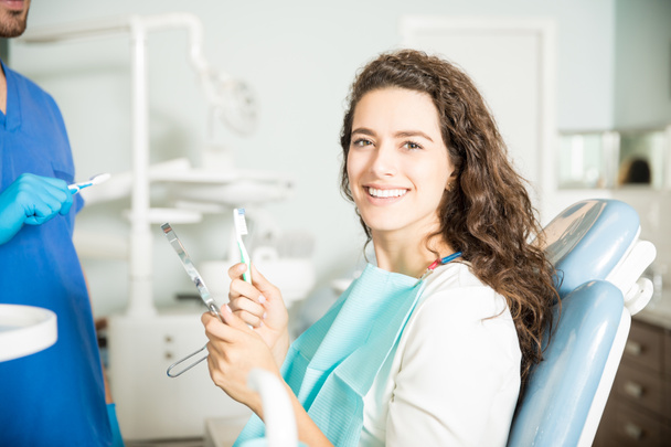 Портрет улыбающейся молодой женщины с зубной щеткой и зубным зеркалом стоматолога в клинике
 - Фото, изображение