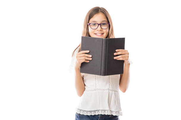 Portrait of little adorable girl wearing white shirt and spectacles holding open book against white background  - Photo, Image