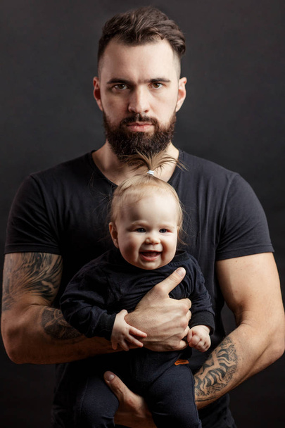 Handsome tattooed beared man holding cute little baby on black background - Photo, Image
