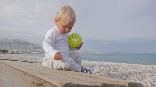 Portret van een blonde baby spelen op de achtergrond van de zee. De baby een gele bal houden met puistjes spelen met kiezels op het strand. - Video