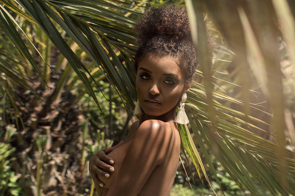 Tropical shoot of beautiful african american woman wearing only earring, posing over palm leaves. - Fotó, kép