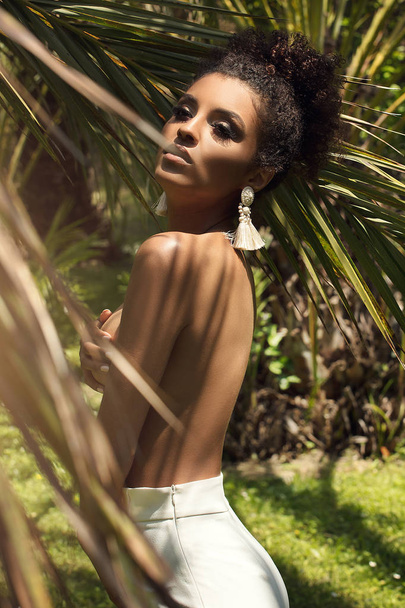 Tropical shoot of beautiful african american woman wearing only earring, posing over palm leaves. - 写真・画像