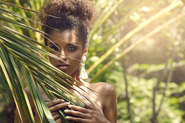 Tropical shoot of beautiful african american woman wearing only earring, posing over palm leaves. - Foto, Imagem