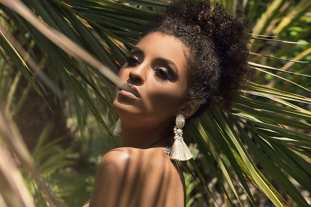 Tropical shoot of beautiful african american woman wearing only earring, posing over palm leaves. - Fotó, kép