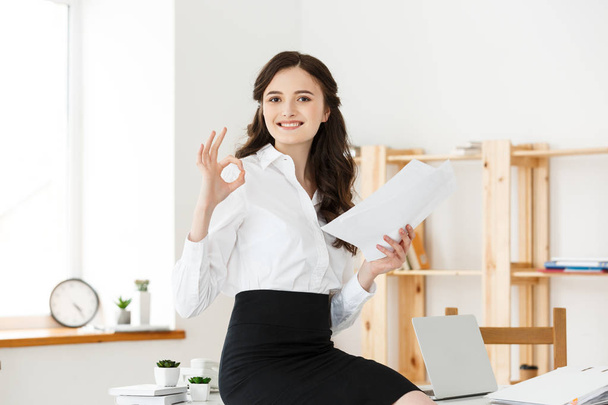 Молодая деловая женщина показывает ОК знак с держателем документа и сидя в современном офисе
 - Фото, изображение