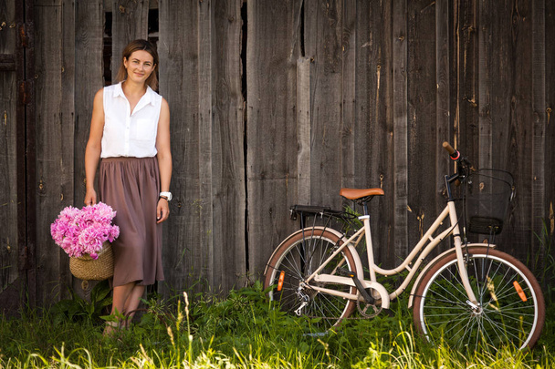Молода темноволоса жінка в білій сорочці в бежевій спідниці тримає красивий букет з рожевих півоній у плетеному кошику. поруч бежевий ретро велосипед на фоні дерев'яного паркану
 - Фото, зображення