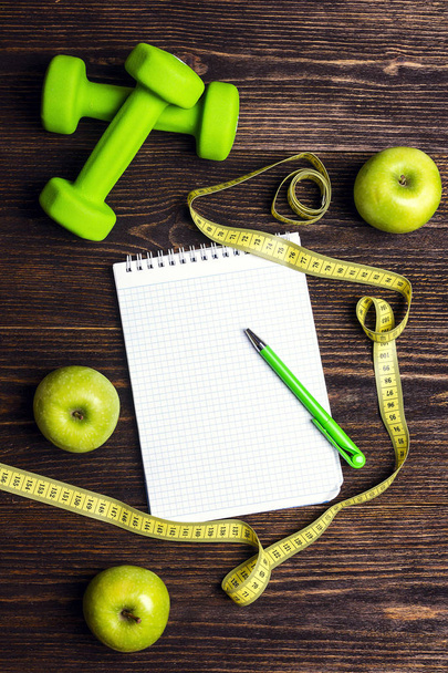 Open blank notepad with green dumbbells and green apples with measuring tape and copy space on wooden background. Sport, fitness and diet concept. Top view.  - Foto, immagini