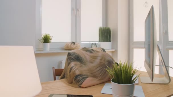 Moe van het wakker worden op het werk. Moe blonde vrouwelijke kantoormedewerker in elegant pak ontspannen liggend op de armen op het bureau dan opstaan geeuwen en beginnen te werken op de computer in de lichte kamer met planten. - Video