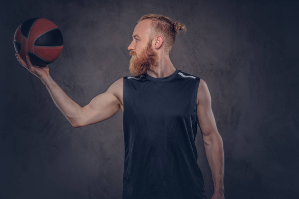 Retrato de un jugador de baloncesto pelirrojo en una ropa deportiva negra sosteniendo la pelota, aislado sobre un fondo de textura oscura
. - Foto, imagen