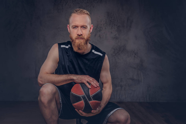 Portréja egy vörös hajú kosárlabda játékos egy gazdaság labdát, elszigetelt texturált sötét háttéren fekete sportruházat. - Fotó, kép