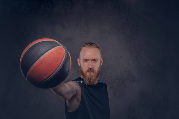 Портрет рыжеволосого баскетболиста в черной спортивной форме, держащего мяч, изолированный на темном текстурированном фоне
. - Фото, изображение