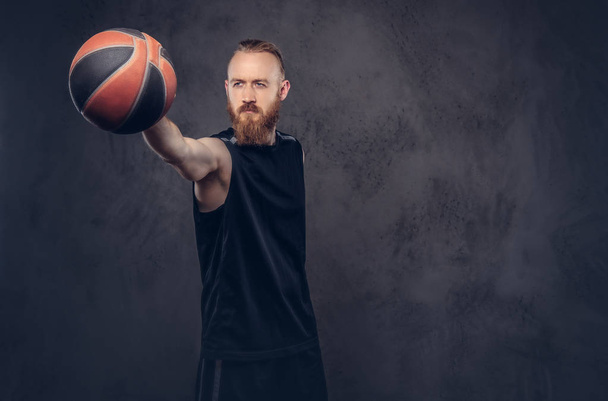 Portréja egy vörös hajú kosárlabda játékos egy gazdaság labdát, elszigetelt texturált sötét háttéren fekete sportruházat. - Fotó, kép