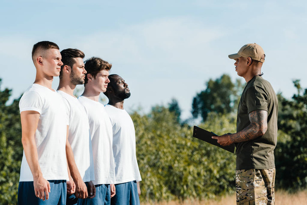 side view of african american tactical instructor with notepad and multiracial young soldiers on range - 写真・画像