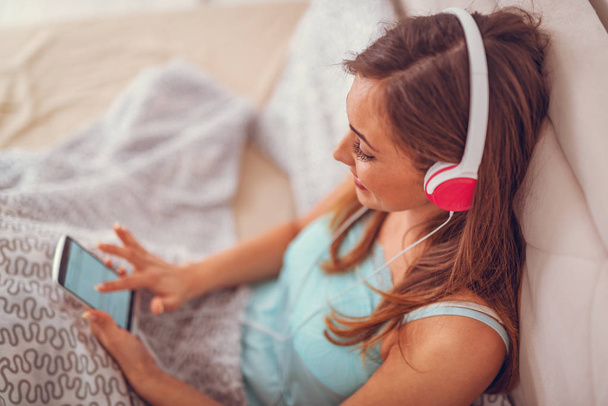 молода усміхнена жінка сидить на ліжку і слухає музику зі смартфона
 - Фото, зображення