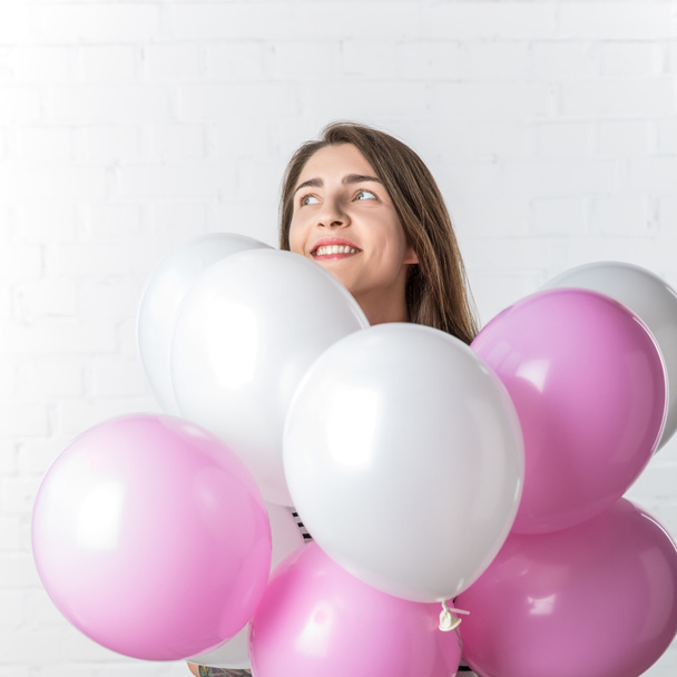 Chica bonita sonriendo sobre un montón de globos en el fondo de la pared de ladrillo blanco
 - Foto, Imagen