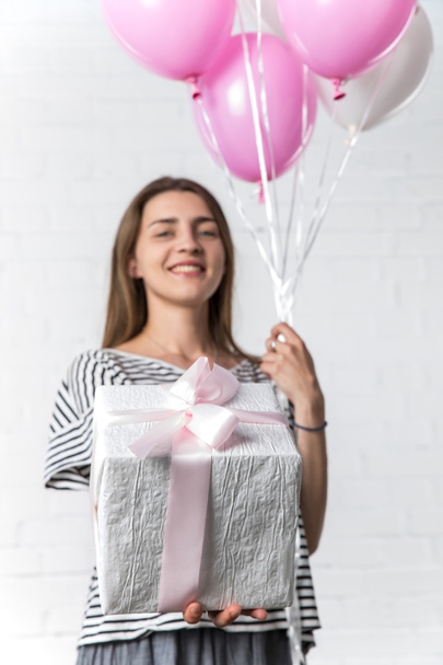 Present and balloons in hands of young girl on white brick wall background - Фото, изображение