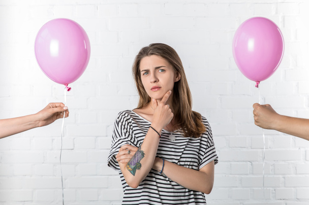 Thoughtful girl standing between hands holding pink balloons on white brick wall background - Foto, immagini
