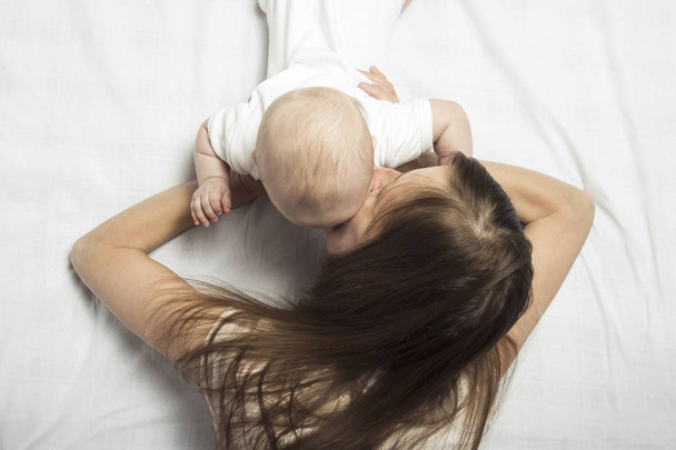 joven madre enseña a gatear un pequeño bebé en la cama con una sábana blanca. Concepto de educación y desarrollo infantil. Piso tendido, vista superior. - Foto, imagen