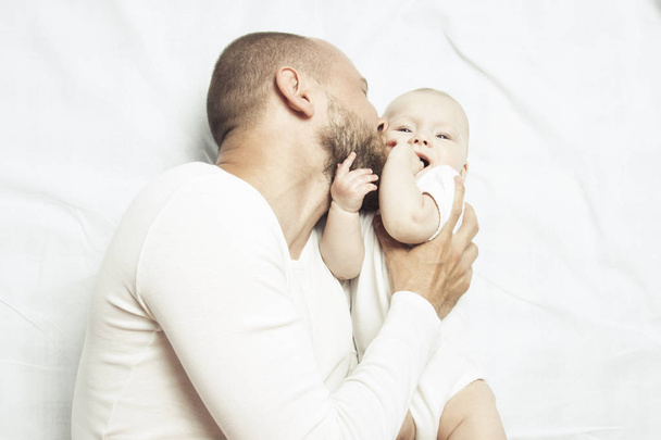Jeune papa jouant avec le petit bébé en vêtements blancs sur un lit avec un drap blanc. Couché plat, vue du dessus
. - Photo, image