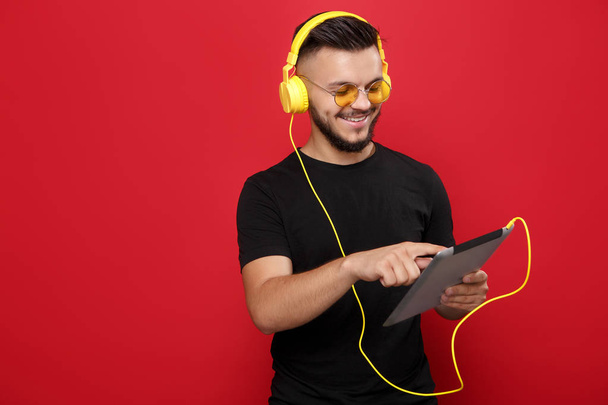 Happy bearded man in yellow sunglasses and black t-shirt listening music and using tablet with happy face on red background. - Foto, Imagem