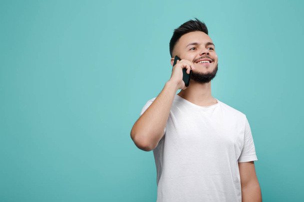 Handsome young bearded hipster in white t-shirt using phone and talking with bright smile. Copy space. - Foto, imagen