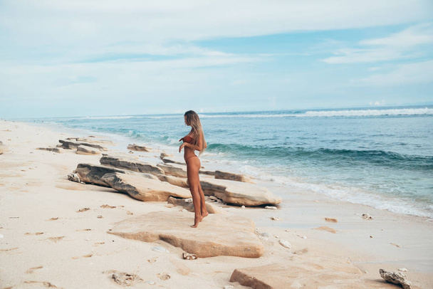 A sexy model in a swimsuit stands in profile and admires the rocks on a deserted white beach. A girl with an even tan and a beautiful body came to relax. - Foto, imagen