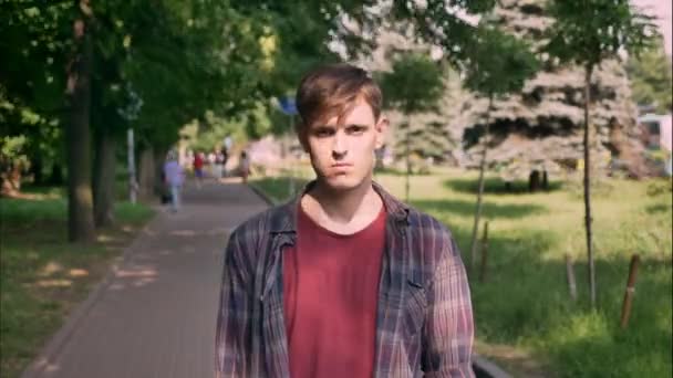 Young serious handsome man is watching at camera in daytime in park in summer, urban concept - Video