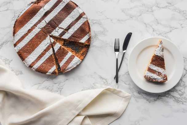 Draufsicht auf köstlichen hausgemachten Kuchen mit Zuckerguss und einem Stück auf Teller mit Gabel und Messer auf Marmoroberfläche - Foto, Bild