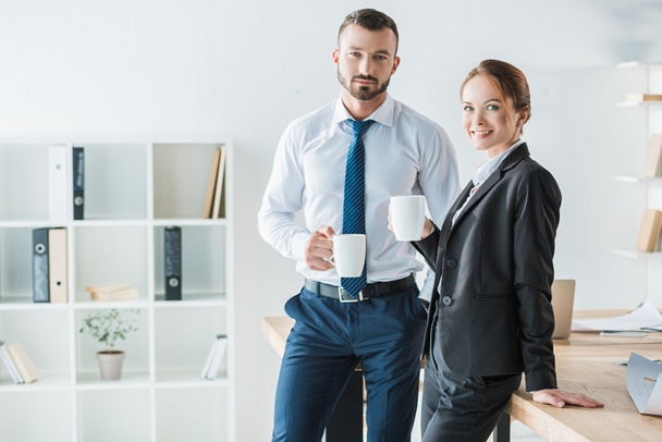 cheerful accountants holding cups with coffee and looking at camera in office - Foto, Imagen