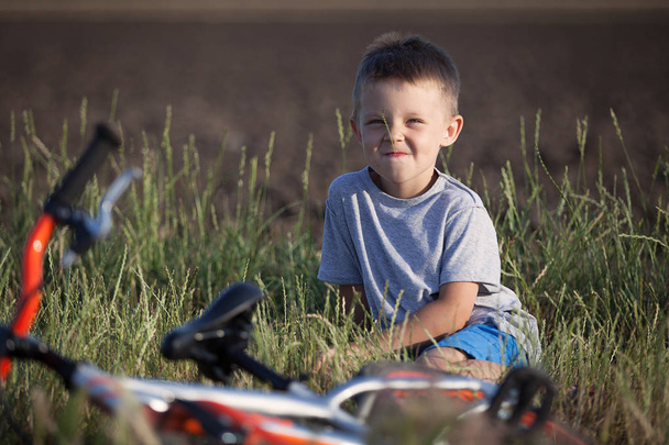 Krásný chlapec sedí v trávě. Vedle něj leží jeho kolo. Sportovní kluk na procházku. - Fotografie, Obrázek