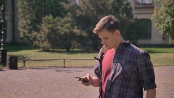 Young handsome man is walking through park in daytime in summer, tapping on smartphone, communication concept - Metraje, vídeo