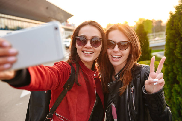 Image closeup of two young european women wearing sunglasses taking selfie photo on smartphone while posing near airport waiting for flight or after departure. Air travel concept - Foto, Imagem