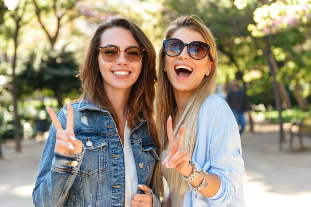 Image of two amazing happy women friends outdoors showing peace gesture looking camera. - Фото, изображение
