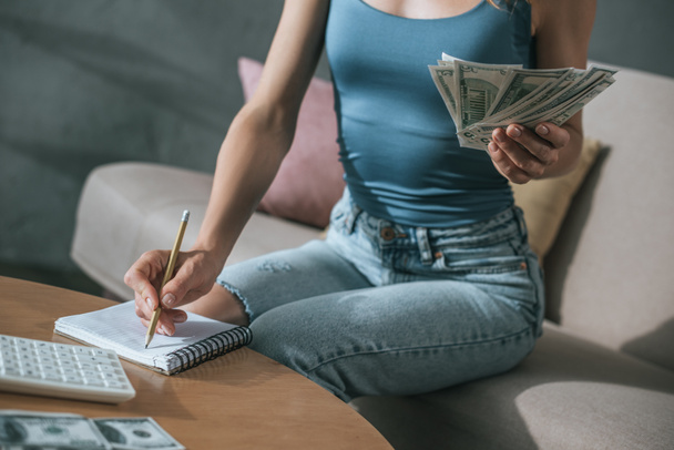 cropped image of woman planning budget at home, holding dollars and writing something to notebook - Fotó, kép