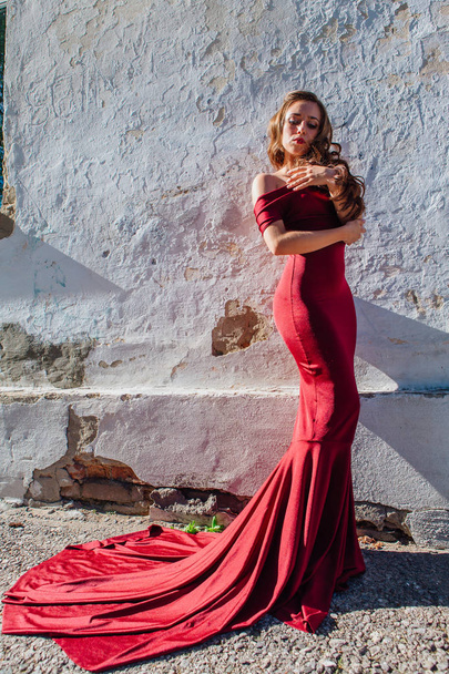 Beautiful young woman in a long red evening gown with a train standing next to the old wall. Copy space - Фото, изображение