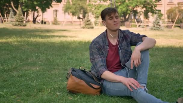 Young handsome man is sitting on grass in park, listening music with earphones, relax concept - Felvétel, videó