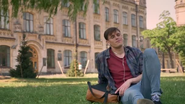 Young handsome man is sitting on grass in park, listening music with earphones, relax concept, building on background - Felvétel, videó