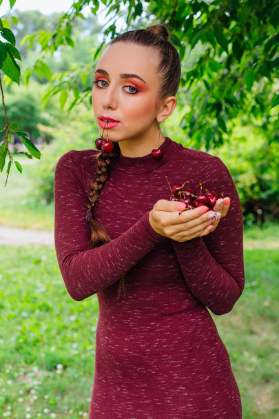 Beautiful young woman with long braid and cherry earrings holding cherry in hands and one cherry in mouth - Foto, Imagem