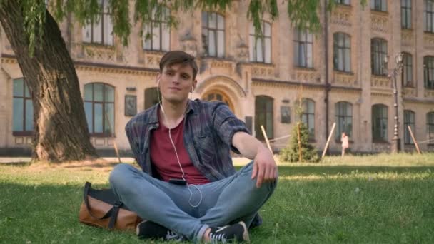 Young handsome man is sitting on lawn in park, listening music with earphones, relax concept, building on background - Кадры, видео