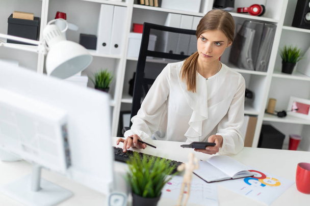 Uma jovem está sentada em uma mesa no escritório, segurando uma calculadora na mão e digitando no teclado
. - Foto, Imagem