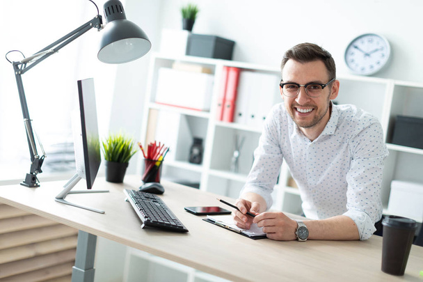 Ein junger Mann mit Brille steht neben einem Tisch im Büro und hält einen Bleistift in der Hand. - Foto, Bild