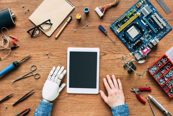 zugeschnittenes Bild eines Elektronikers mit Roboterhand am Tisch mit digitalem Tablet, umgeben von Werkzeugen - Foto, Bild