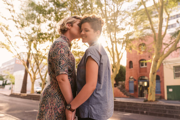 Uśmiechający się młoda kobieta, całował w policzek przez jej miłości dziewczyny podziwiając w słoneczny dzień razem w mieście - Zdjęcie, obraz