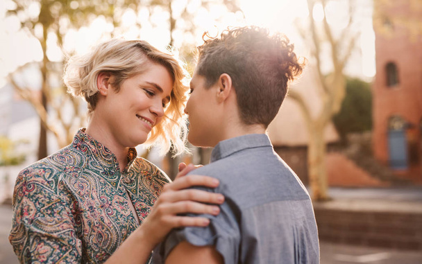 Smiling young lesbian couple standing arm in arm on a sunny street looking into each other's eyes  - Foto, Bild