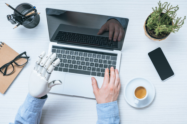 обрізане зображення бізнесмена з протезною рукою, використовуючи ноутбук за столом з горшковою рослиною і чашкою кави
  - Фото, зображення