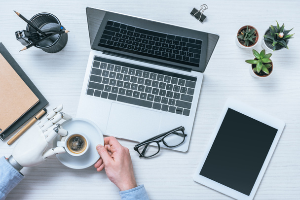 обрезанный образ бизнесмена с протезной рукой, пьющего кофе за столом с ноутбуком, планшетом и очками
  - Фото, изображение