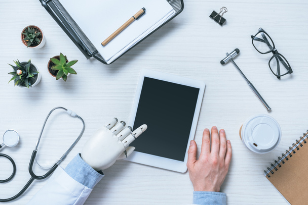 abgeschnittenes Bild eines männlichen Arztes mit prothetischem Arm mittels digitalem Tablet mit leerem Bildschirm am Tisch mit medizinischen Werkzeugen  - Foto, Bild