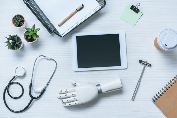 Ansicht des Arztarbeitsplatzes mit Armprothese, Stethoskop, Reflexhammer und digitalem Tablet mit leerem Bildschirm auf dem Tisch  - Foto, Bild