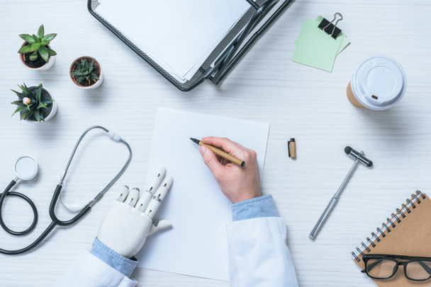 oříznutý obraz mužského lékaře s protézu psaní na čistý papír u stolu s reflexní kladivo, stetoskop a šálek kávy  - Fotografie, Obrázek